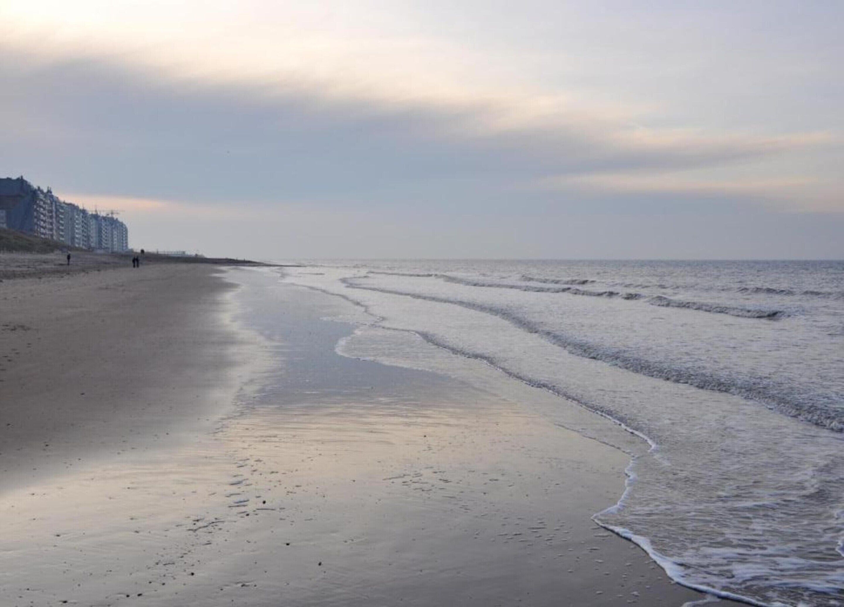 Ein Strand mit ruhiger See, einige Spaziergänger und Gebäude im Hintergrund.