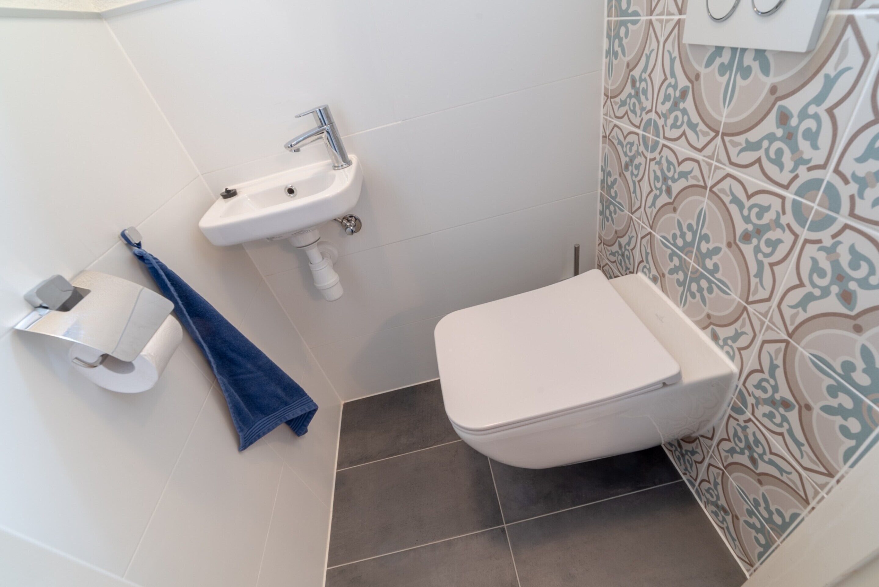 Casa de banho com uma sanita, lavatório branco e paredes com azulejos estampados.