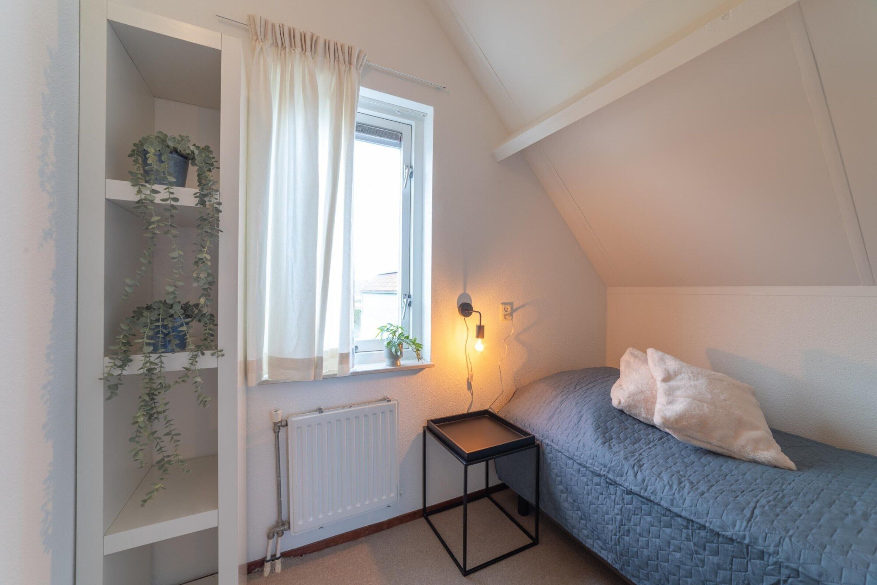 Een slaapkamer met een schuine kap, een raam met een witte gordijn, een nachtkastje met een lamp, een bed met een blauwe quilt en een radiator.
