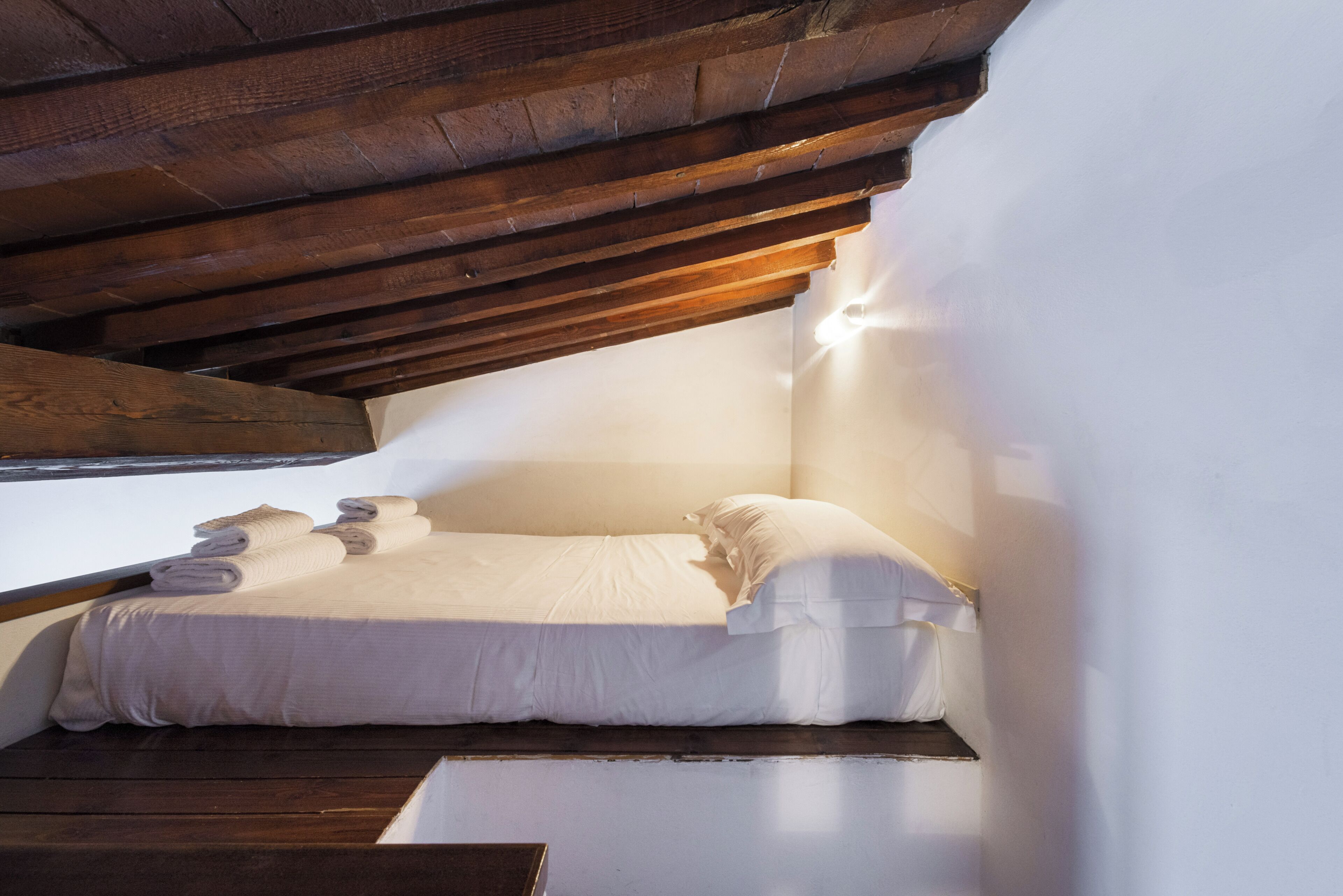 A small, cozy room with a single bed, wooden beams overhead, and a window providing natural light.