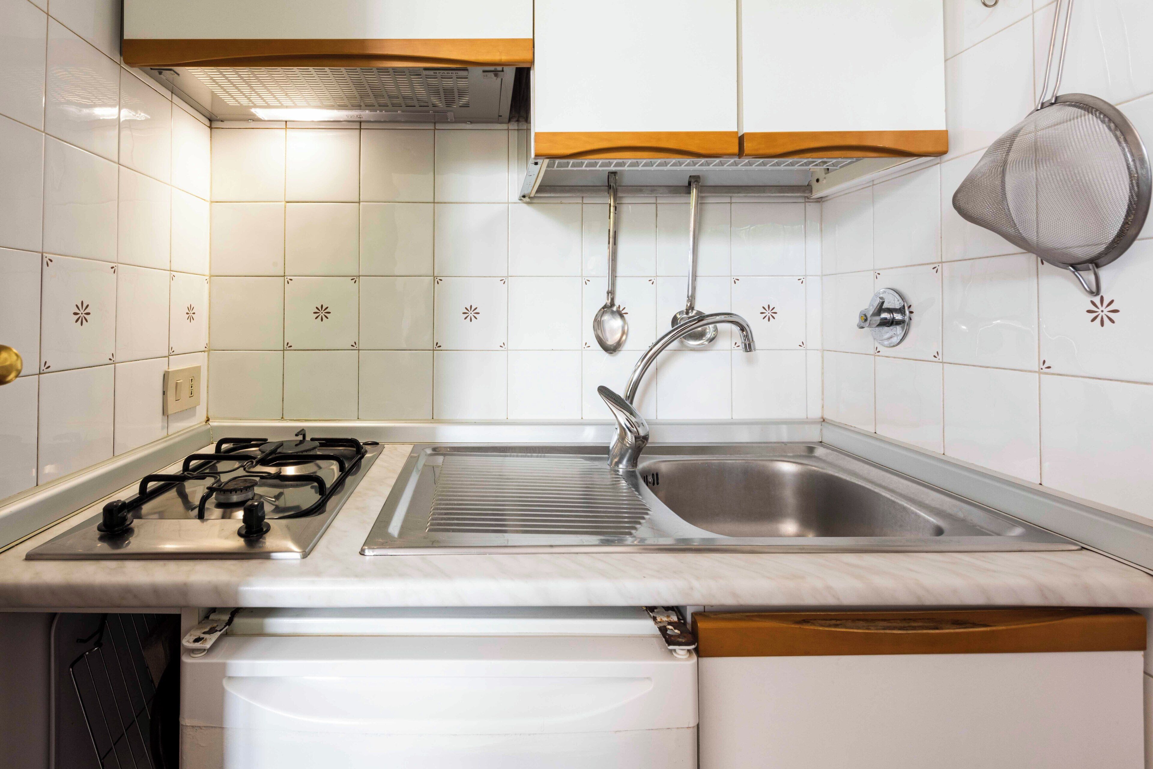 Une cuisine avec des carreaux blancs, un évier en acier inoxydable, une cuisinière à gaz et des armoires en bois.
