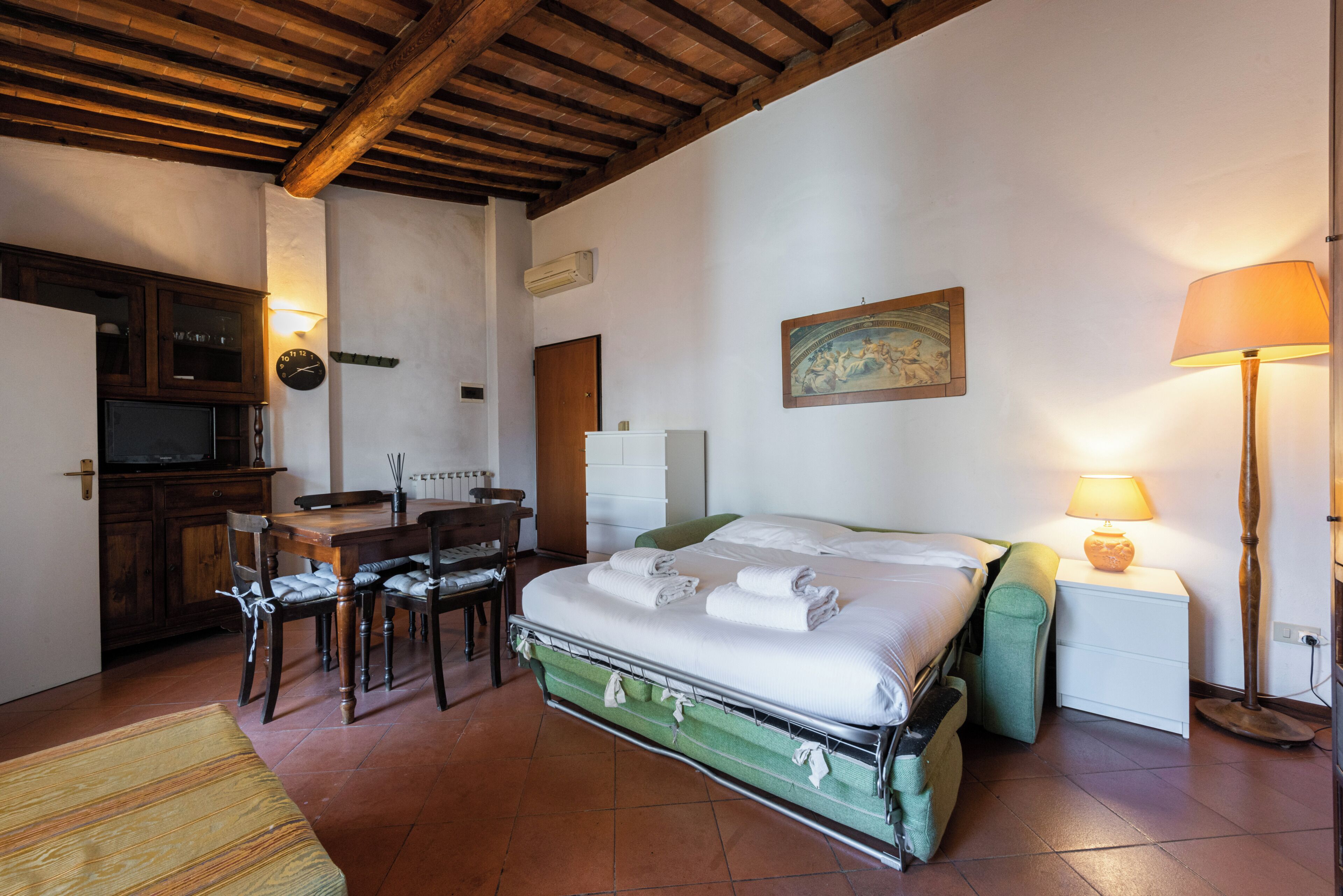 A room with a bed, a table, chairs, a lamp, and a painting on the wall.