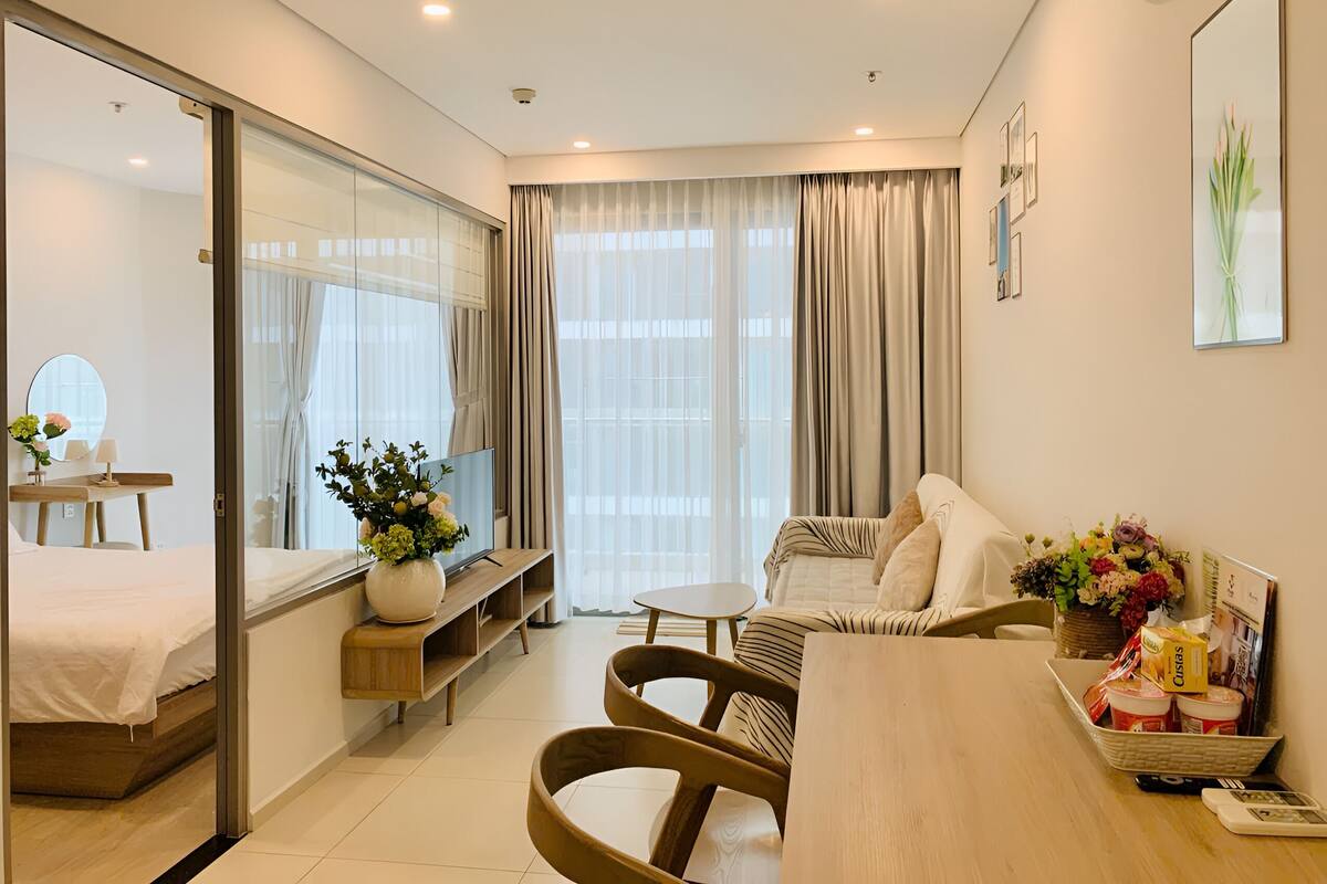 Ein modernes Hotelzimmer mit Bett, einer Sitzecke inklusive Sofa und Sesseln, einem Esstisch mit einer Platte mit Speisen und einem großen Fenster mit Vorhängen.