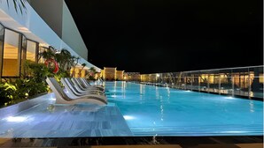 Vista nocturna de una piscina con sillones reclinables y un edificio moderno con barandillas de vidrio.