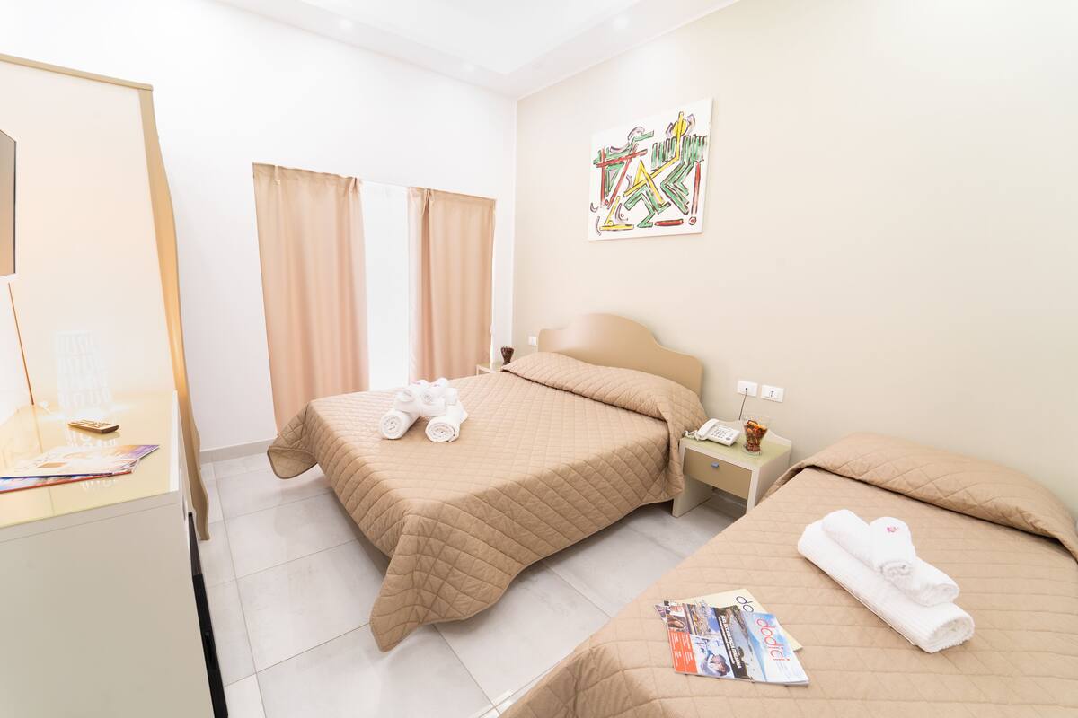 A hotel room with two beds, a desk, a mirror, a window with curtains, and a wall art piece.