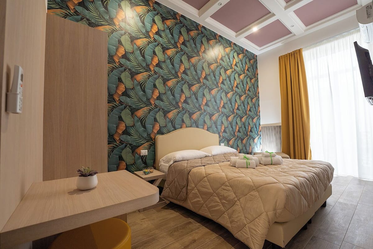 A hotel room with a bed, a desk, a small table with a plant, and a wall with a leaf pattern.