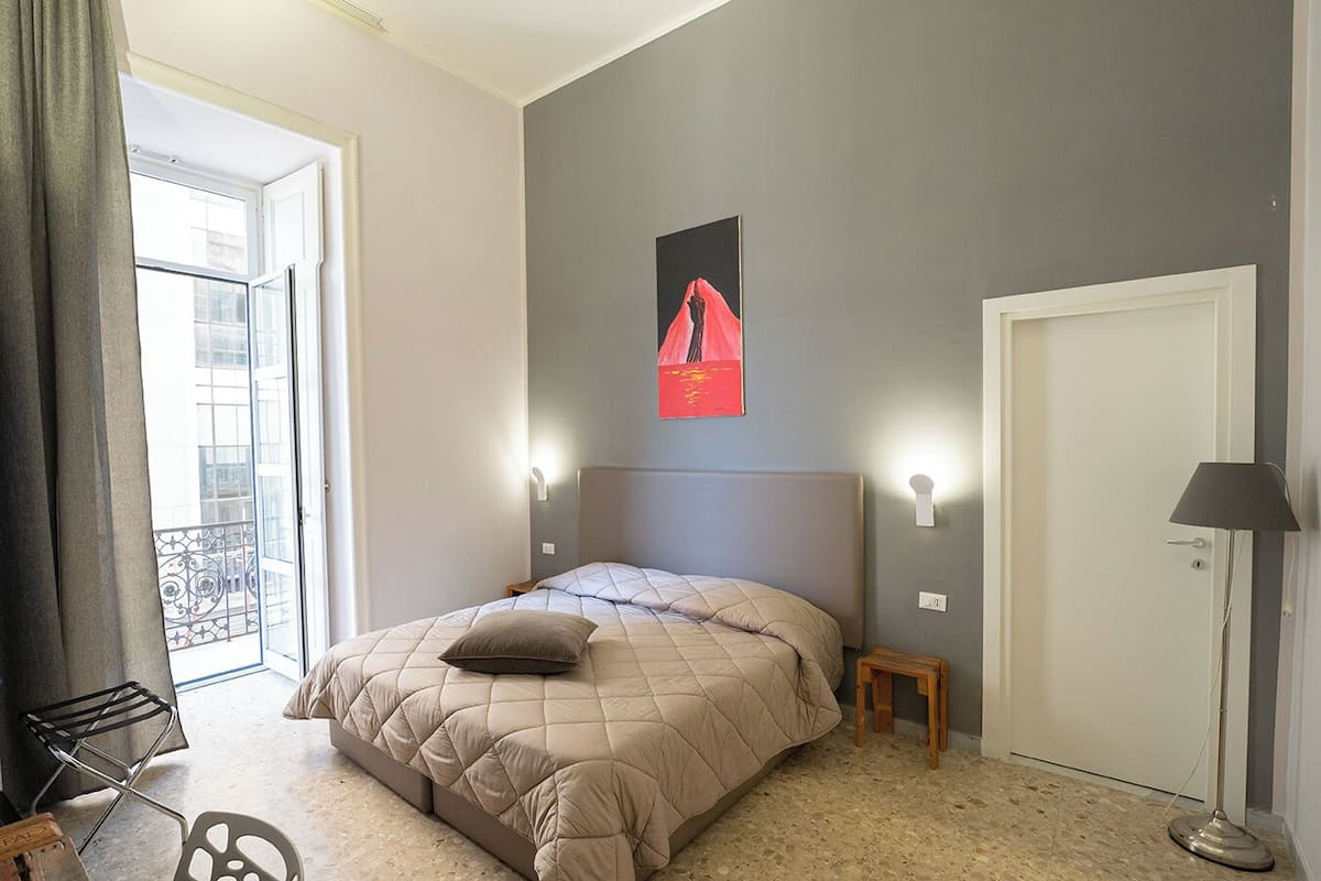 A bedroom with a bed, a small bedside table, a lamp, a door, and a window with a view of buildings.