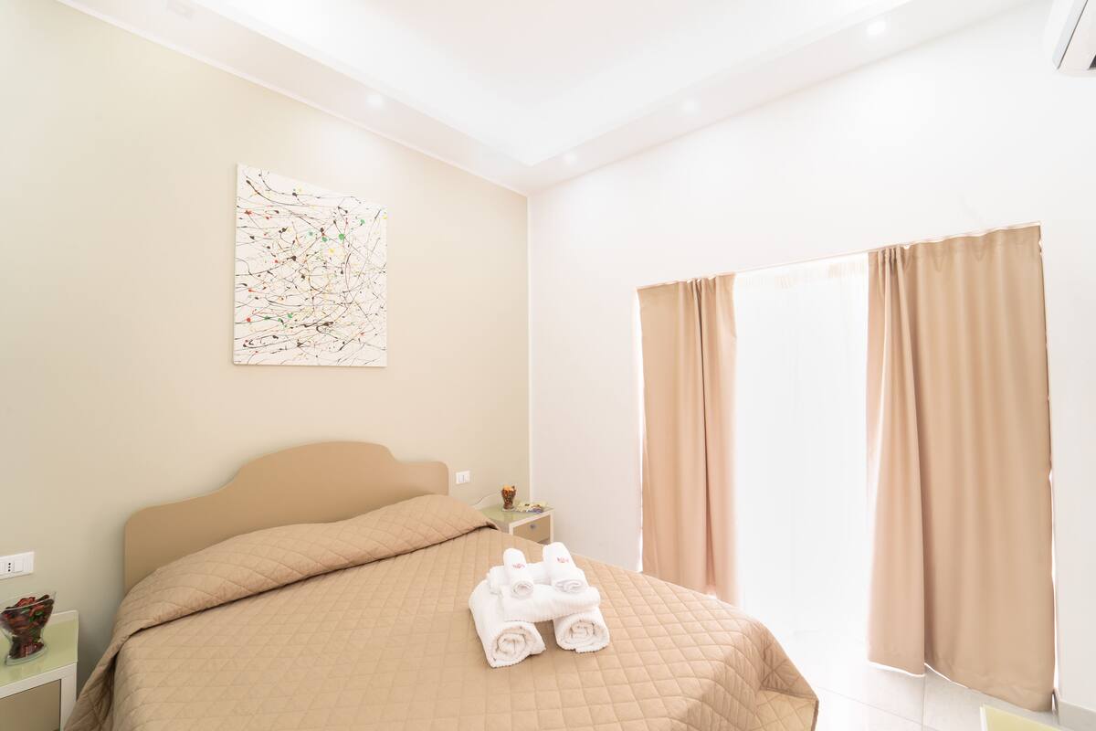 A hotel room with a bed, a nightstand, a painting, and a window with curtains.