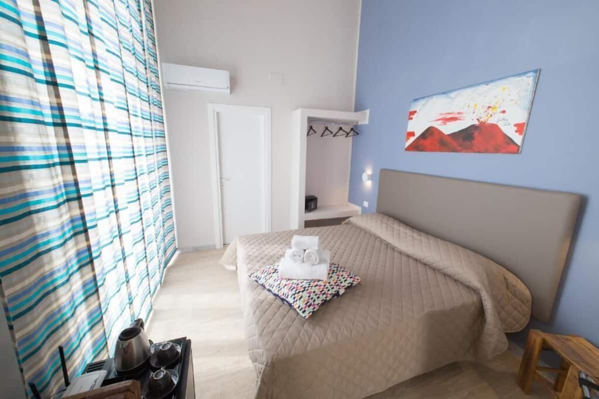 A bedroom with a bed, a bedside table, a chair, a window with curtains, a painting on the wall, and a door.