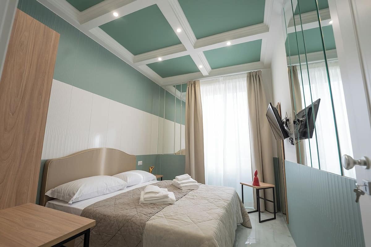 A hotel room with a bed, a bedside table, a TV mounted on the wall, and a window with curtains.