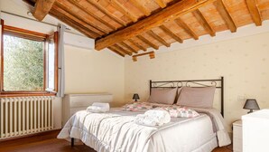A bedroom with a wooden ceiling, a radiator, a bed with pillows, a bedside table, and a window with a view of greenery.