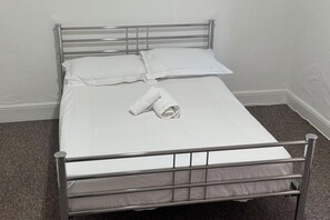 A metal-framed bed with white bedding and a rolled towel, set against a plain wall and carpeted floor.