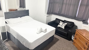 Un dormitorio con cama, un sofá negro con cojín estampado de hojas, un aparador de madera y una ventana con cortinas.