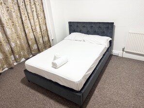 A neatly made bed with a dark headboard in a room with a carpeted floor and a radiator.