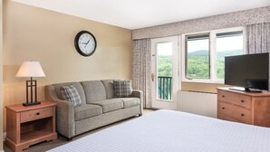A hotel room with a bed, a sofa, a TV, a clock, and a window with a view of greenery. - King Room | Ski-In Resort Stay (West Dover)
