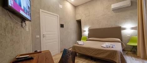 A hotel room with a bed, a desk with a chair, a TV mounted on the wall, and an air conditioning unit.