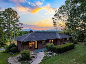 A single-story brick house with a dark roof, surrounded by well-maintained lawns and trees, set against a backdrop of mountains and a colorful sky. - Romantic Asheville Fall Mountain Getaway with Stargazing Hot Tub & Warm Fire Pit (Candler)