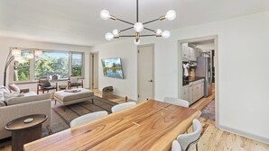 A modern living room with a wooden dining table, sofas, and a kitchen in the background.