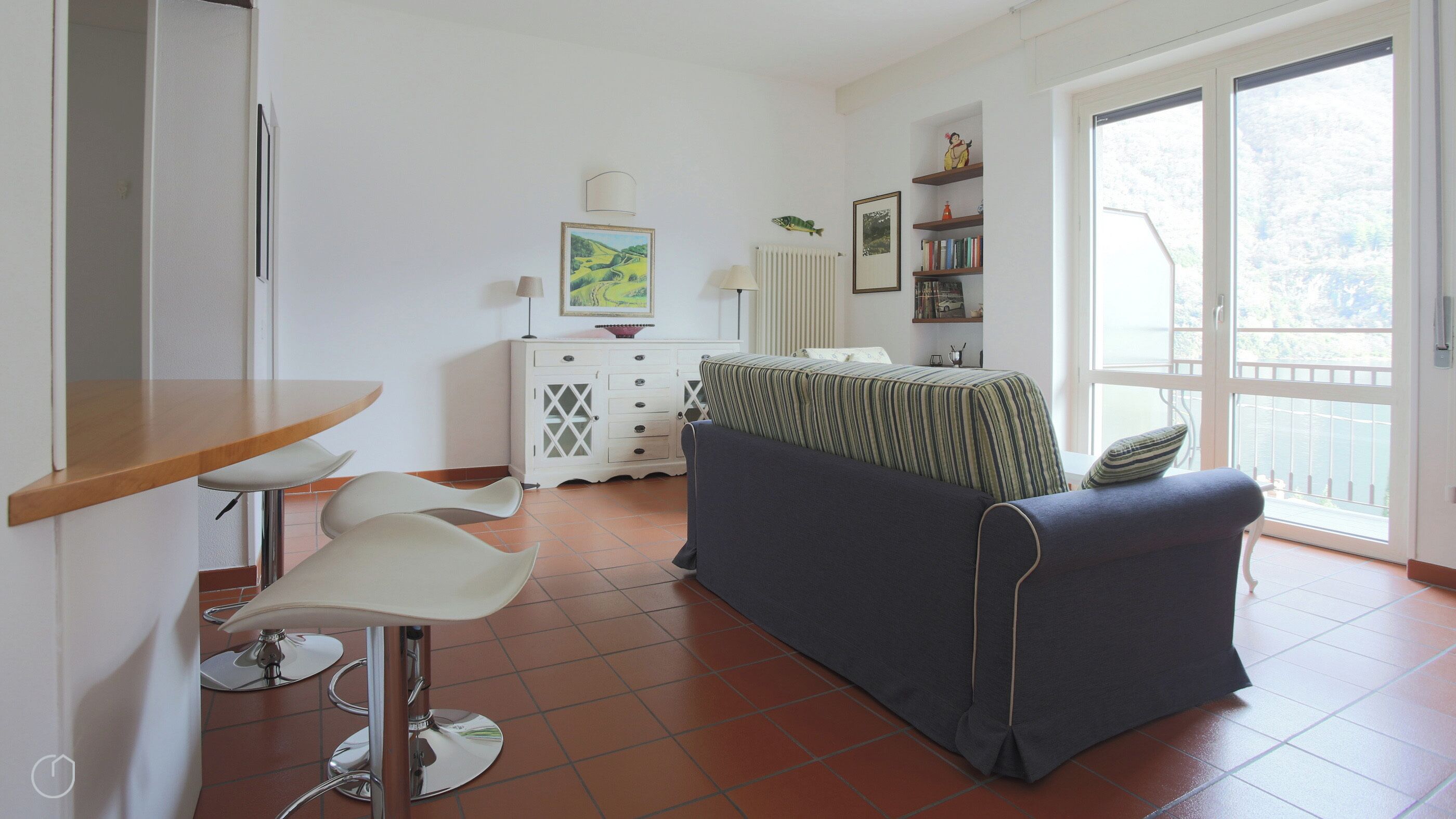 A compact living space with a kitchenette, a bed with striped bedding, a dining area with a wooden table and white chairs, and a view through a large window.