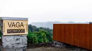 A wooden sign with "VAGA ROWOOD" on a stone base, set against a backdrop of greenery and distant mountains. - Vaga Rowood by WanderEase (Vagamon)