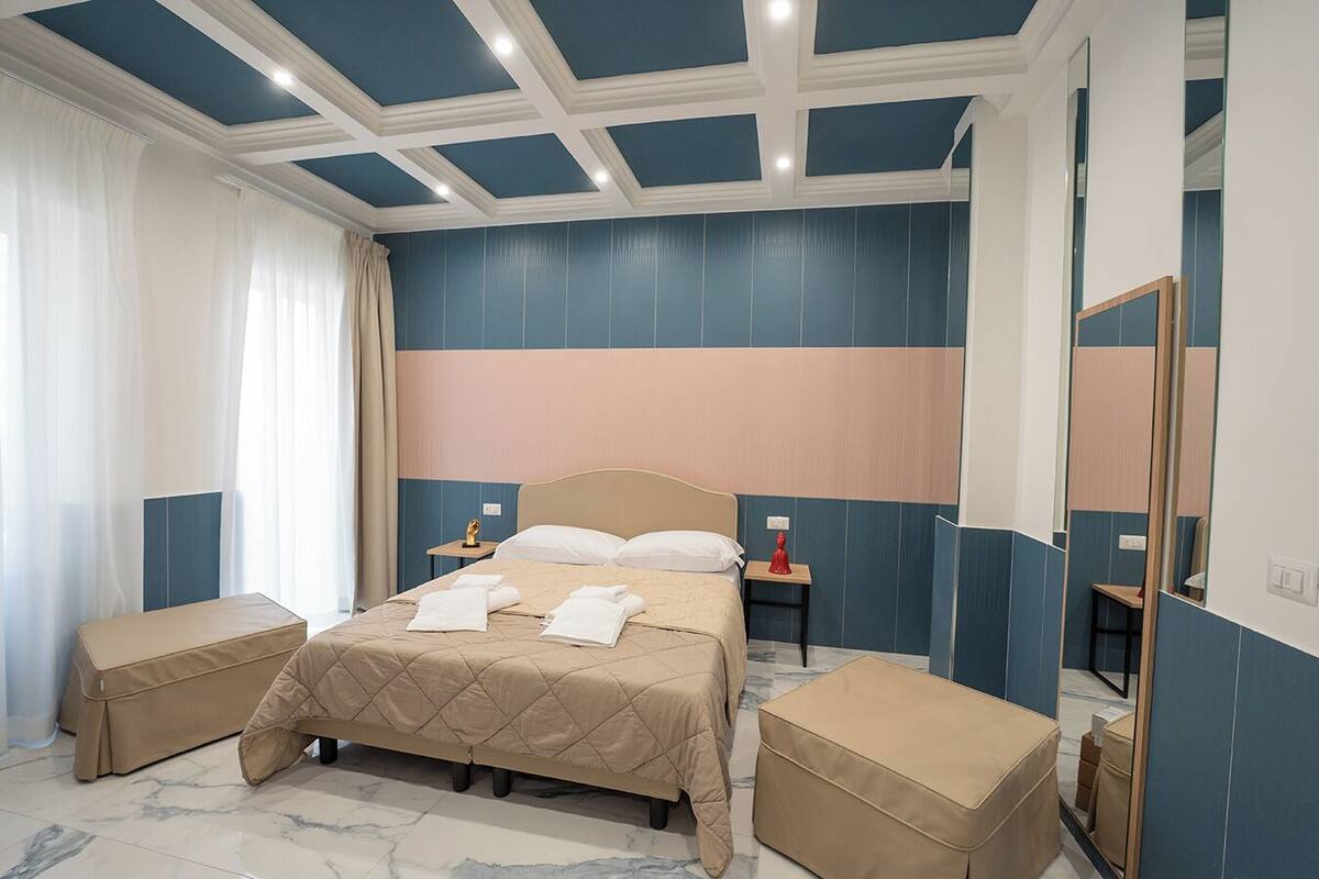 A hotel room with a large bed, two bedside tables, a mirror, and a window with sheer curtains.