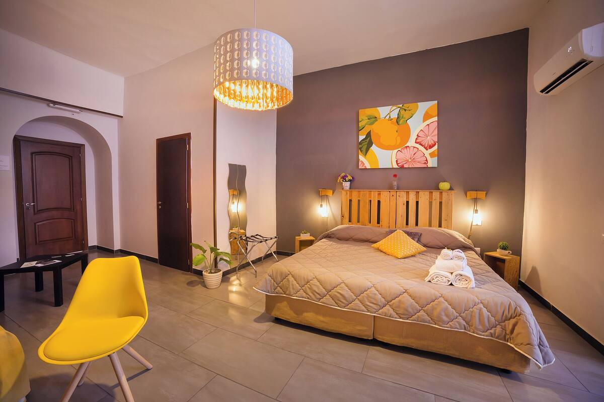 A modern bedroom with a large bed, a bright yellow chair, a hanging light fixture, and a wall-mounted painting.