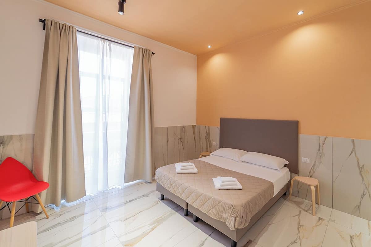 A hotel room with a bed, a red chair, and a window with curtains.