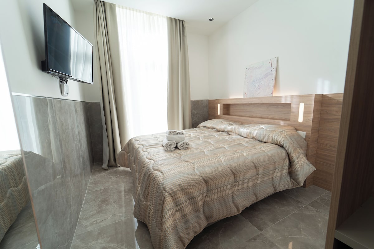 A modern hotel room with a large bed, a flat-screen TV mounted on the wall, and a large window with curtains.