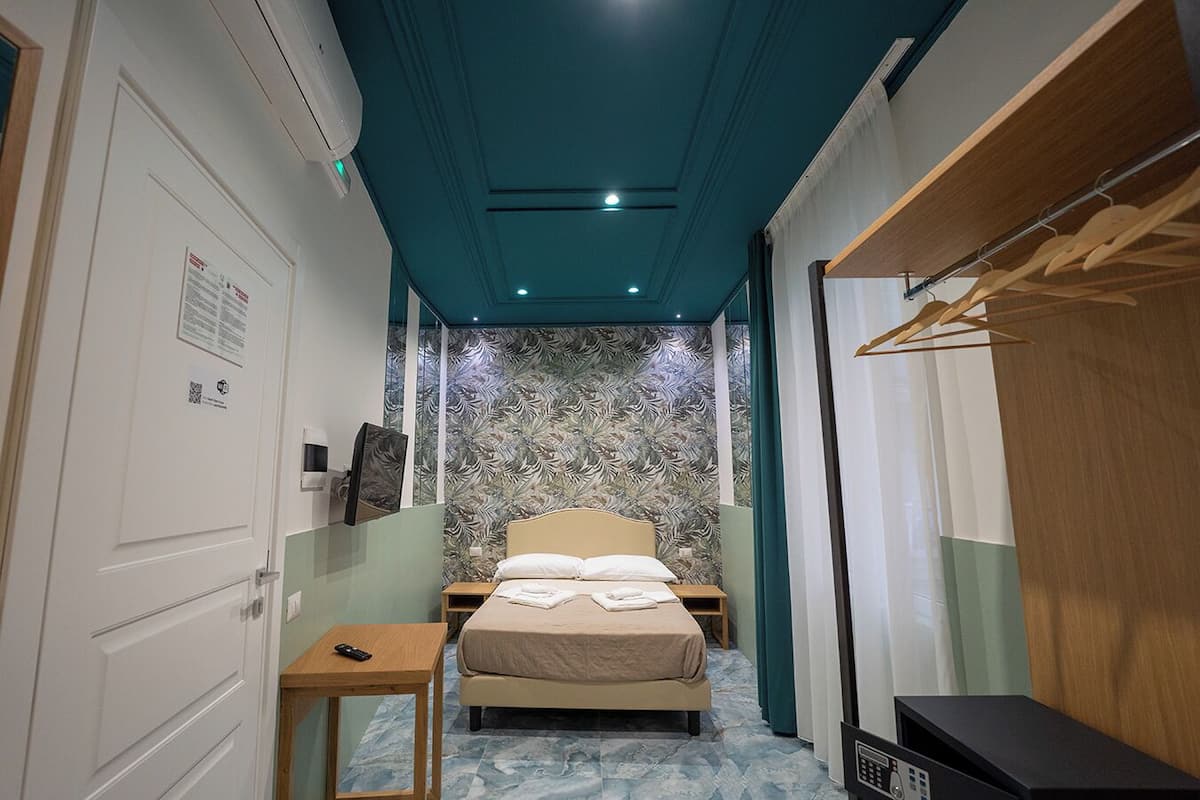 A narrow hotel room with a bed, a bedside table, a small table, a wardrobe, and a mirror.