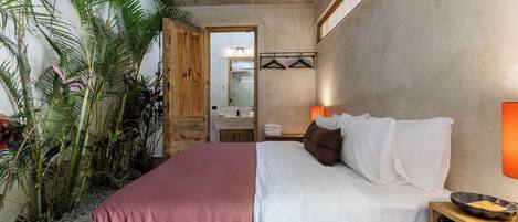 Un dormitorio con cama, mesita de noche y espejo. Se ven plantas y una puerta de madera al fondo.