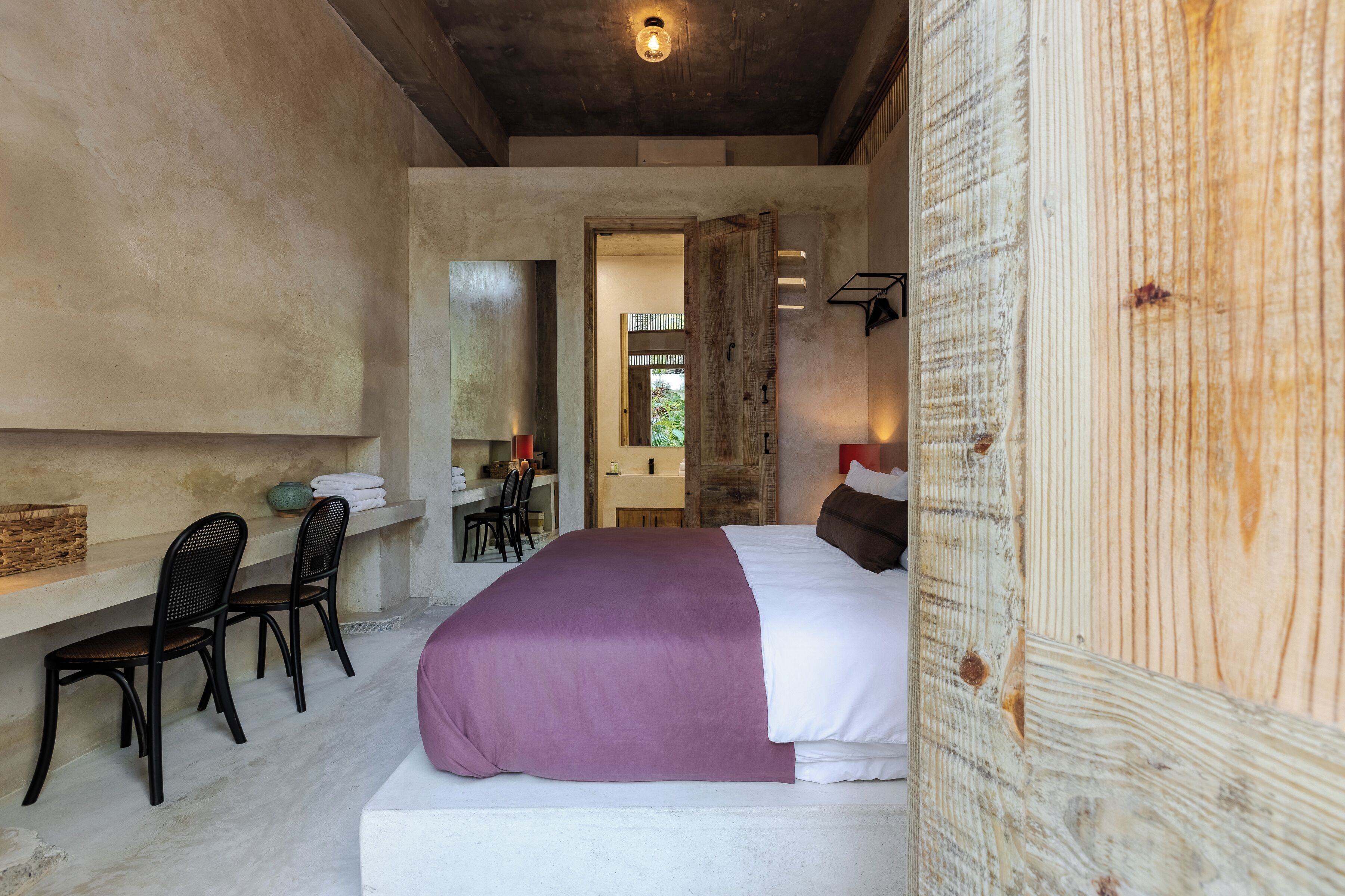 Ein Schlafzimmer mit einem großen Bett, einem hölzernen Kopfteil, einer Bank mit Stühlen und Blick auf ein Badezimmer und einen Garten.