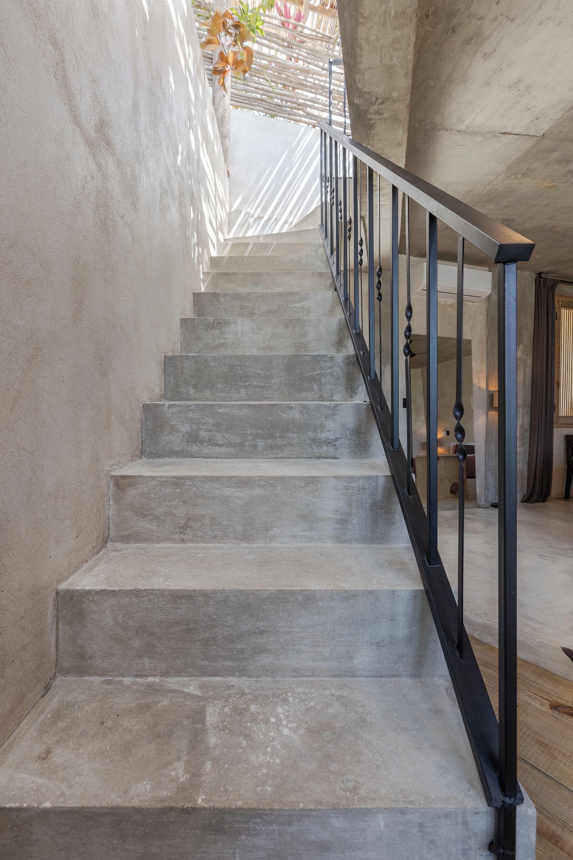 Una escalera de concreto con barandales de metal que lleva a una habitación con un ventanal grande y una planta.