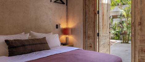 A bedroom with a bed, pillows, a bedside lamp, and a view of a garden through an open door.