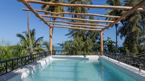 Una piscina trasparente con un pergolato in legno sopra, circondata da palme e altra vegetazione.