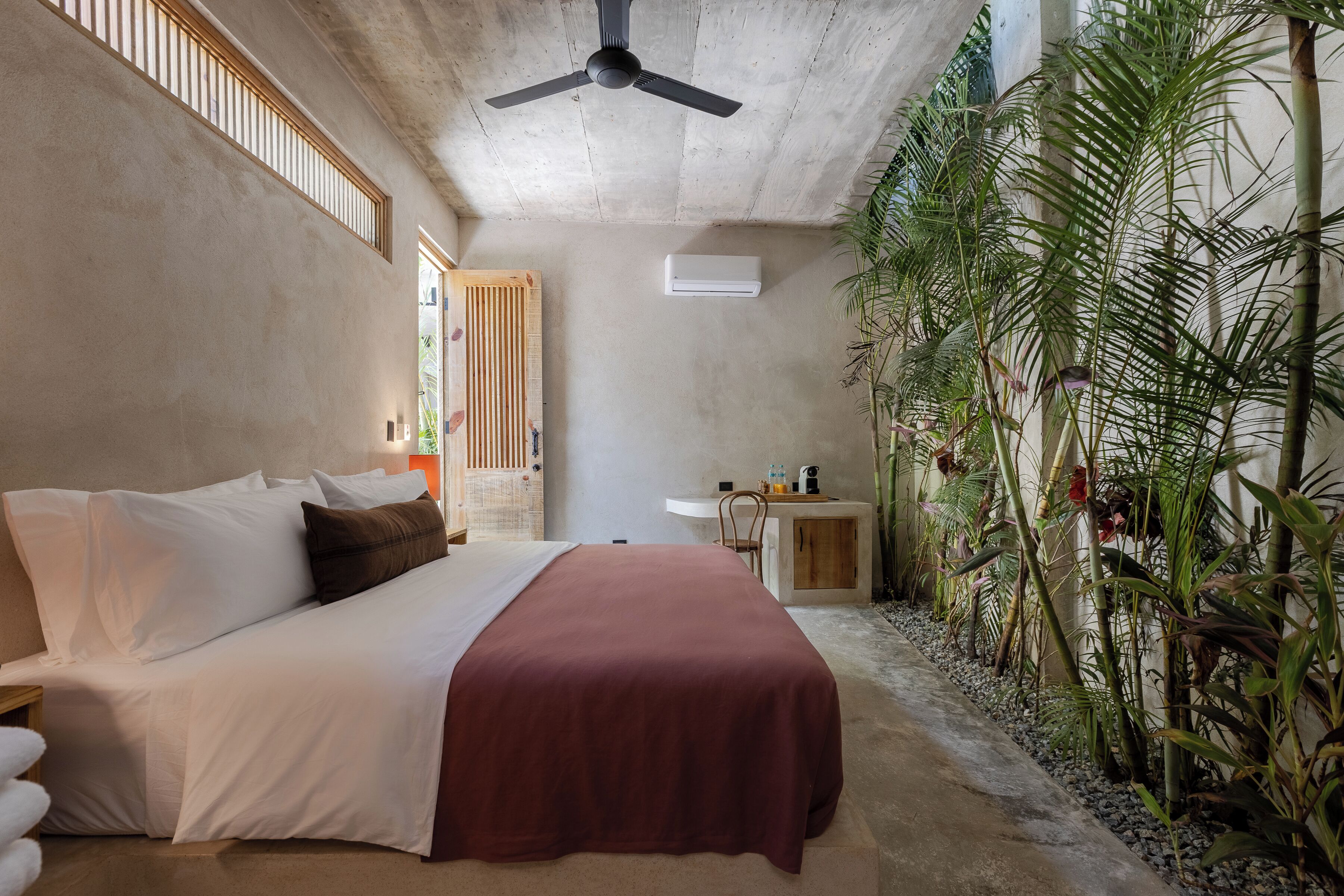 A bedroom with a bed, a ceiling fan, an air conditioning unit, a small desk, and a plant area.