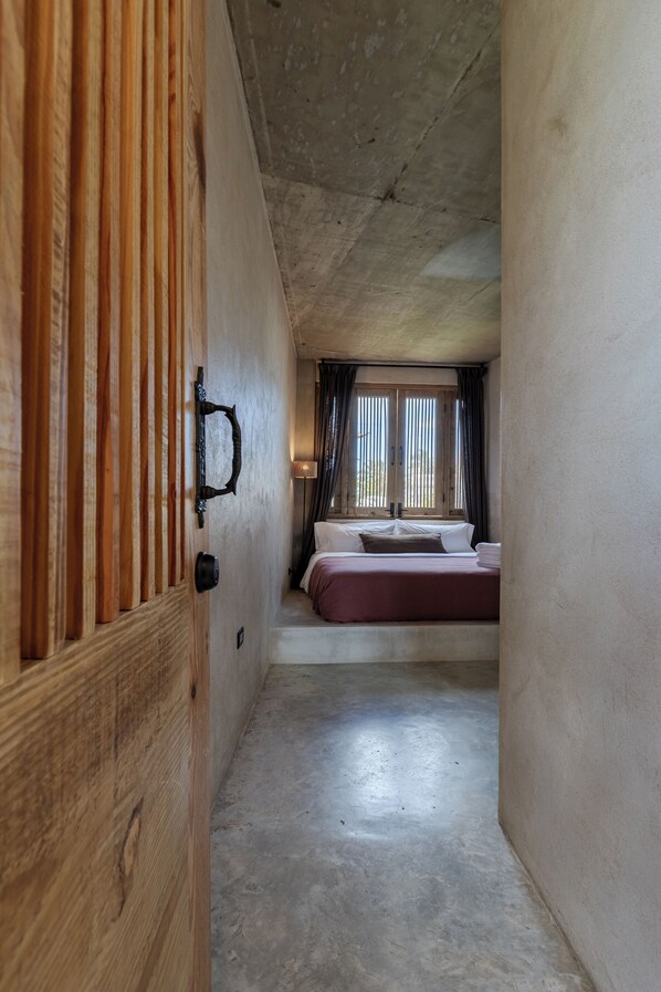 A narrow hallway leading to a bedroom with a bed, a window with curtains, and a wooden door.