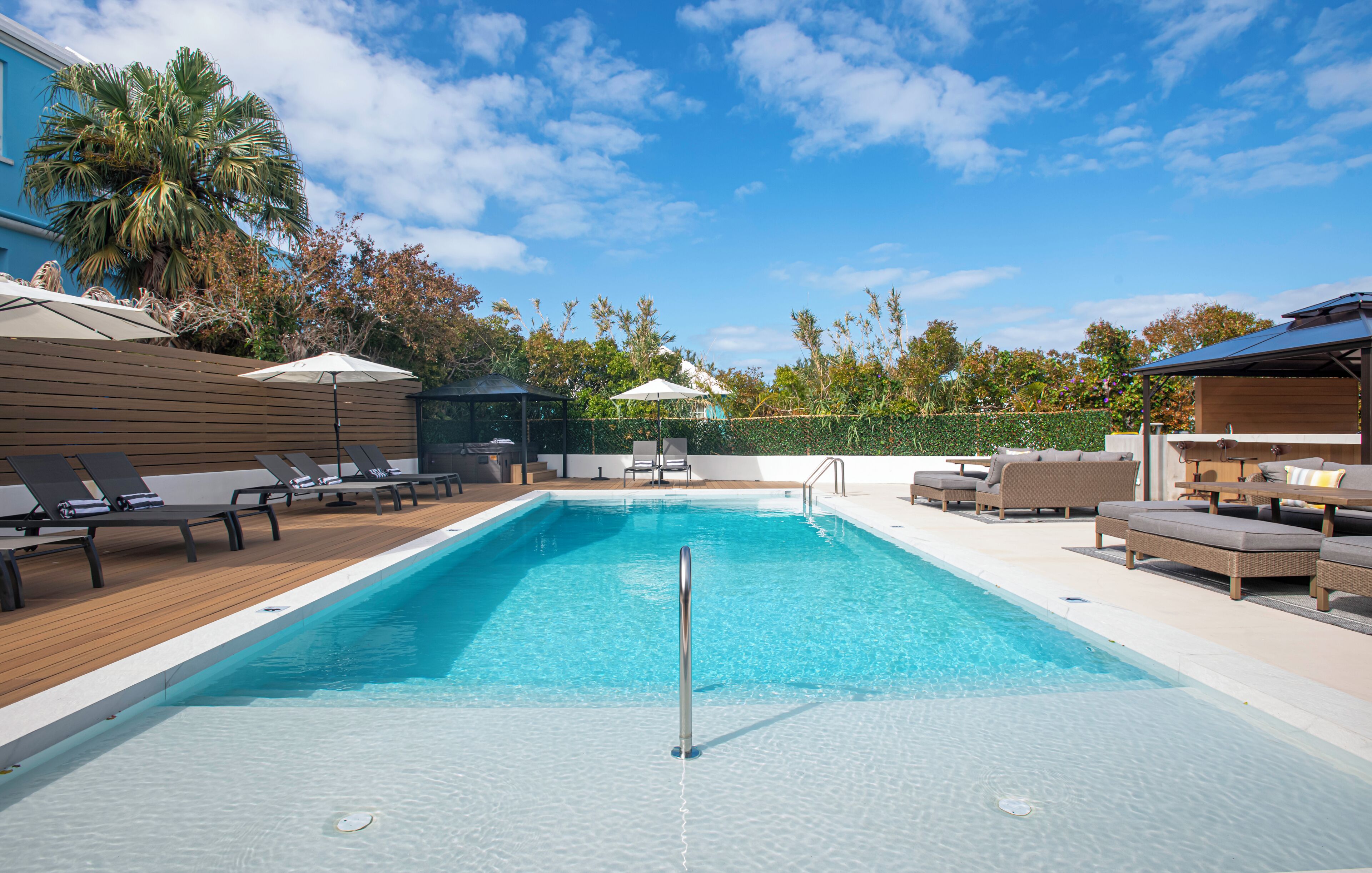 Una piscina rectangular con muelle de madera, sillas de descanso, sombrillas y una zona de estar cubierta.