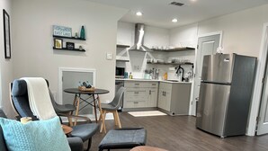 Una sala de estar moderna con un sofá gris, una mesa de centro de madera y un comedor con una mesa redonda y sillas. Se ve una cocina en el fondo, con gabinetes blancos, un refrigerador de acero inoxidable y un horno empotrado.
