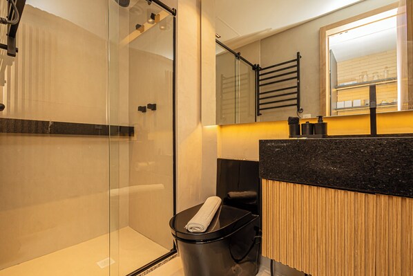 Un baño moderno con cabina de ducha de vidrio, un cubo de basura negro con una toalla, un tocador de madera y un espejo sobre el lavabo.