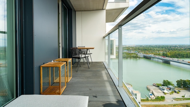 Ein Balkon mit Blick auf einen Fluss und eine Brücke, Holzmöbel und große Fenster.