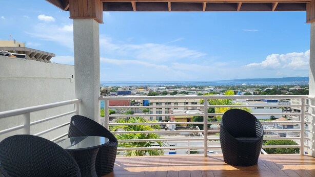 A balcony with wicker chairs and a glass table, overlooking a cityscape with buildings and a distant view of the sea. - 2-bedroom apartment in delightful Kingston with WiFi, fitness room, AC (Kingston)