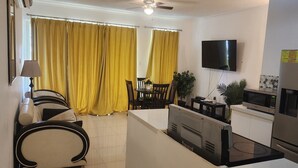 A living room with a sofa, a dining table with chairs, yellow curtains, a TV, a microwave, and a refrigerator. - 2-bedroom apartment in delightful Kingston with WiFi, fitness room, AC (Kingston)