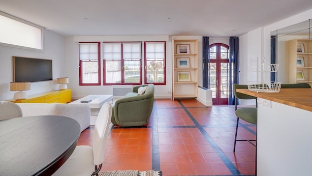 Ett modernt vardagsrum med en soffa, ett soffbord och en tv. Det finns en matplats med ett träbord och stolar. Rummet har stora fönster med röda ramar och en dörr med röd och blå design.