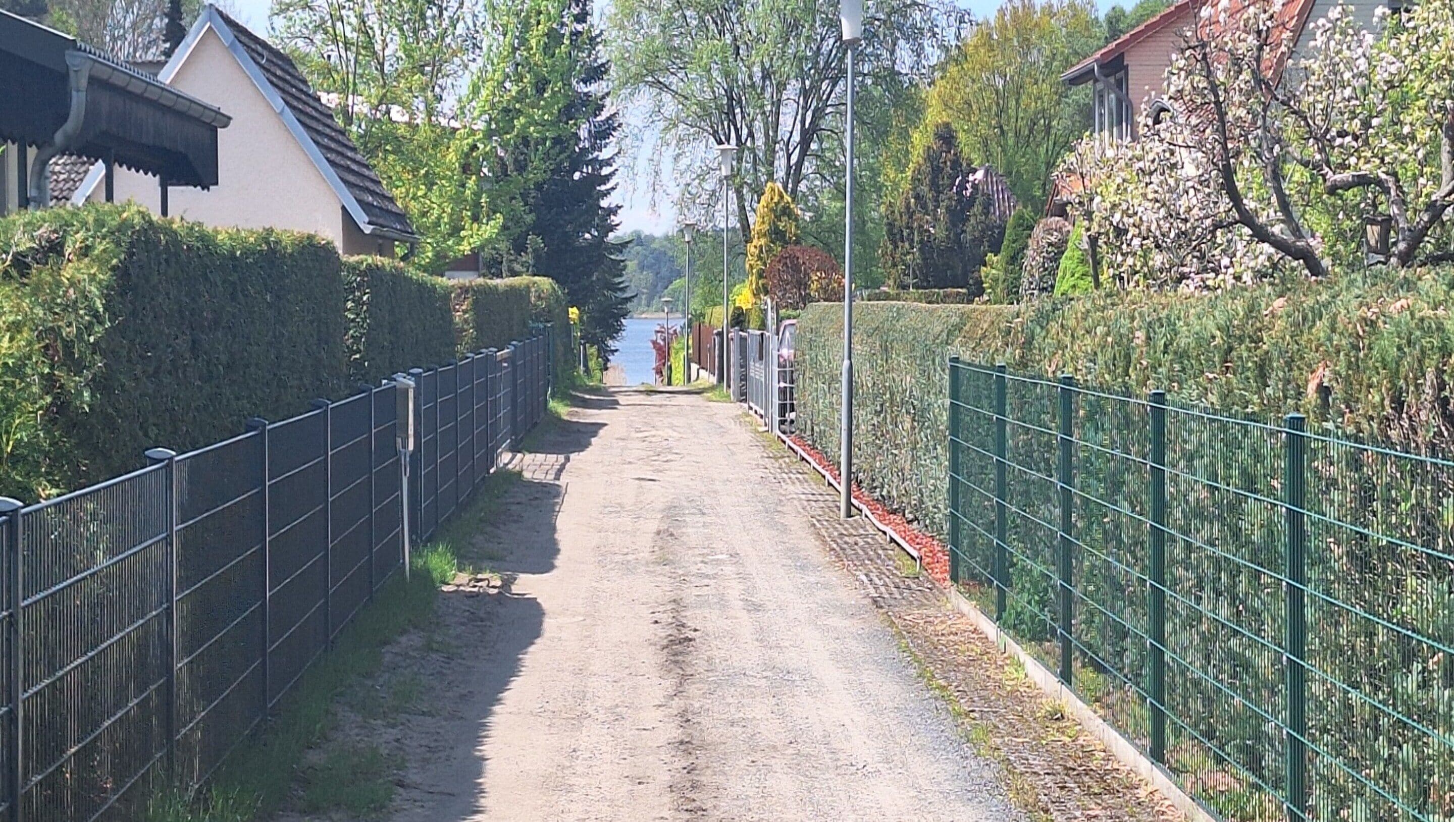 Un sentier étroit, pavé, bordé de clôtures métalliques et de haies, qui mène à un plan d’eau.