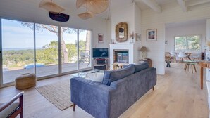 A spacious living room with a large gray sofa, a fireplace, and a television. There are large windows offering a view of the outdoors. - Villa Belvédère: pool, and beach 5 minutes- Pyla (La Teste-de-Buch)