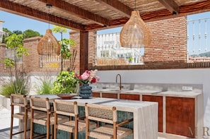 Cocina exterior con barra, sillas de mimbre, pileta y luminarias colgantes.