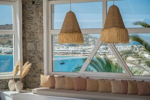 Une pièce avec un mur en pierre, une vue sur une ville côtière, une fenêtre donnant sur la mer et un coin salon avec des coussins.
