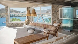 A modern interior with a stone wall, large windows, a wooden table, and a seating area with cushions. - Villa Delta Ornos - MG Villas Mykonos (Mykonos)
