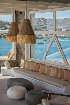 A room with a large window overlooking a body of water, a woven hanging light fixture, a white cushioned bench with pillows, and a stone wall. - Villa Delta Ornos - MG Villas Mykonos (Mykonos)