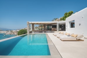 A modern poolside area with a clear view of the sea, a covered seating area, and a white building with large windows.
